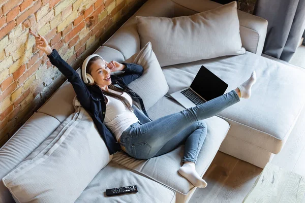 Overhead view of happy girl listening music in wireless headphones near laptop with blank screen and remote controller — Stock Photo