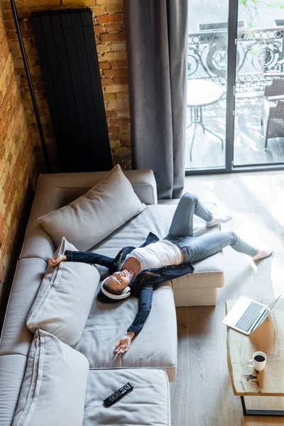 Overhead view of happy woman in wireless headphones listening music near laptop and remote controller while lying on sofa — Stock Photo