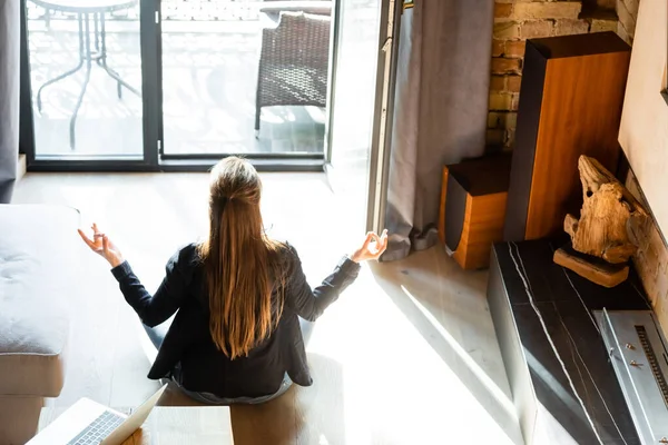 Back view of young woman practicing yoga and sitting in lotus pose near laptop — Stock Photo