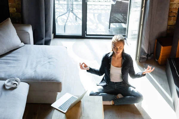 Attractive young woman with closed eyes sitting in lotus pose near laptop — Stock Photo