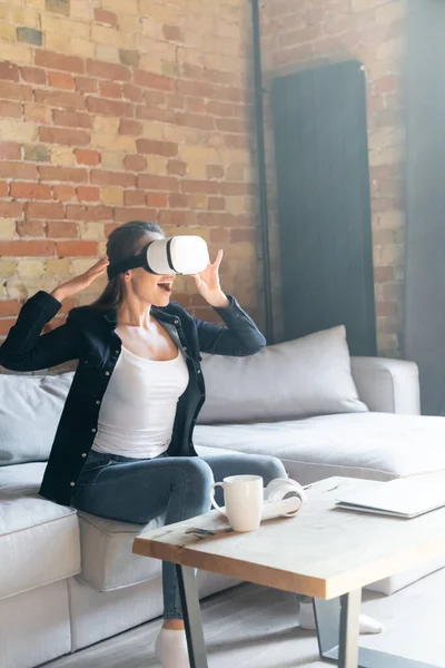 Aufgeregte junge Frau berührt Virtual-Reality-Headset im Sitzen auf dem Sofa — Stockfoto