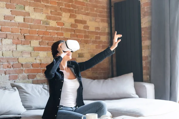 Mujer joven y feliz tocando los auriculares de realidad virtual y el gesto mientras está sentado en el sofá - foto de stock