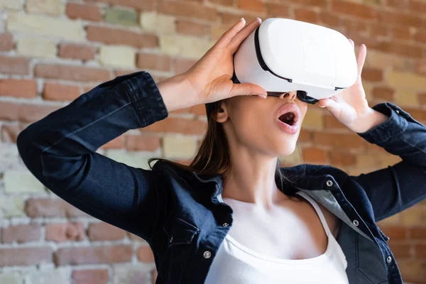 Schockierte junge Frau berührt Virtual-Reality-Headset gegen Ziegelwand — Stockfoto