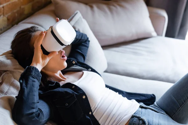Удивлённая девушка, касающаяся гарнитуры виртуальной реальности и лежащая на диване — стоковое фото