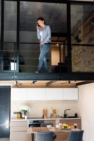 Молодая женщина разговаривает по смартфону, стоя на втором этаже в двухуровневой квартире — стоковое фото