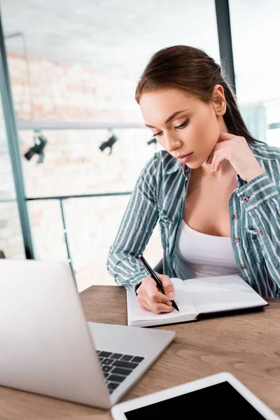 Selective focus of beautiful woman writing in notebook near laptop and digital tablet with blank screen, online study concept — Stock Photo