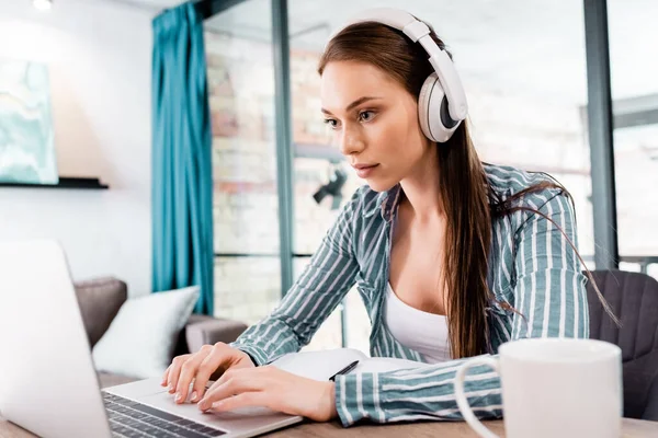 Selective focus of beautiful freelancer in wireless headphones using laptop near cup — Stock Photo