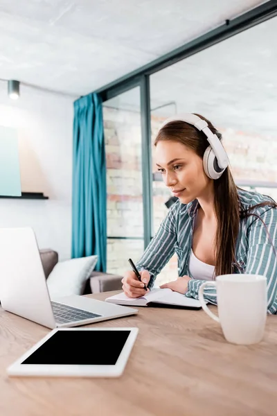 Selective focus of girl in wireless headphones writing in notebook near laptop and digital tablet with blank screen, online study concept — Stock Photo