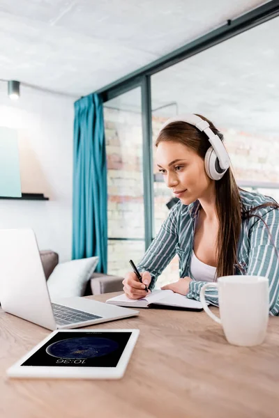 KYIV, UKRAINE - APRIL 29, 2020: selective focus of girl in wireless headphones writing in notebook near laptop and ipad, online study concept — Stock Photo