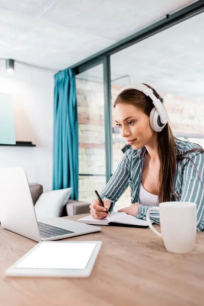 Selective focus of girl in wireless headphones writing in notebook near laptop and digital tablet with white screen, online study concept — Stock Photo