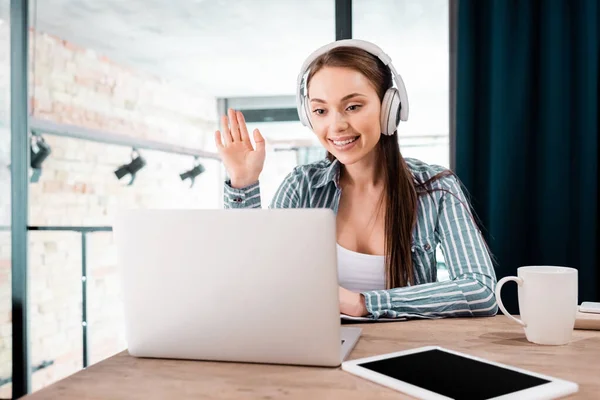 Selective focus of girl in wireless headphones waving hand near laptop and digital tablet with blank screen — Stock Photo