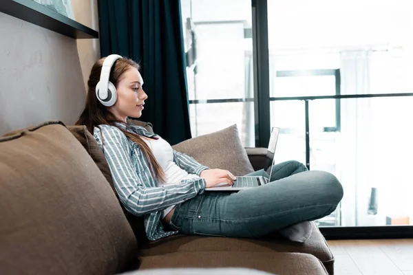 Vista lateral do freelancer atraente em fones de ouvido sem fio usando laptop na sala de estar — Fotografia de Stock