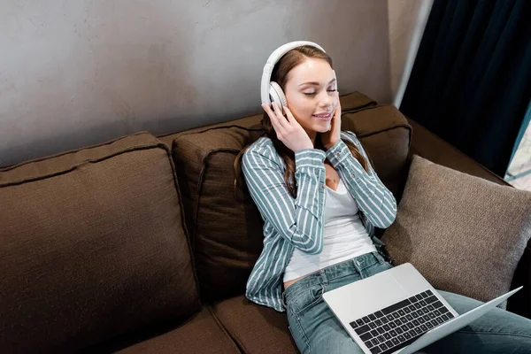 Glückliche Freiberuflerin berührt drahtlose Kopfhörer in der Nähe von Laptop im Wohnzimmer — Stockfoto