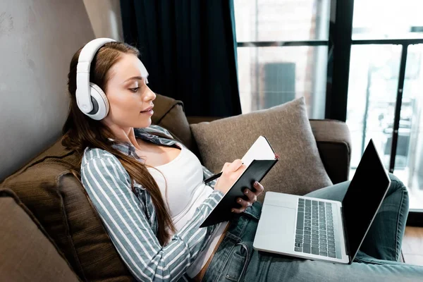 Attraente donna in cuffie wireless scrittura in notebook vicino al computer portatile con schermo bianco, concetto di studio online — Foto stock