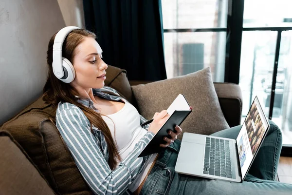 KYIV, UKRAINE - APRIL 29, 2020: attractive woman in wireless headphones writing in notebook near laptop with depositphotos website, online study concept — Stock Photo
