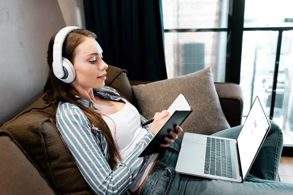 KYIV, UCRANIA - 29 DE ABRIL DE 2020: mujer atractiva en la escritura de auriculares inalámbricos en el portátil cerca de la computadora portátil con el sitio web de Google, concepto de estudio en línea - foto de stock