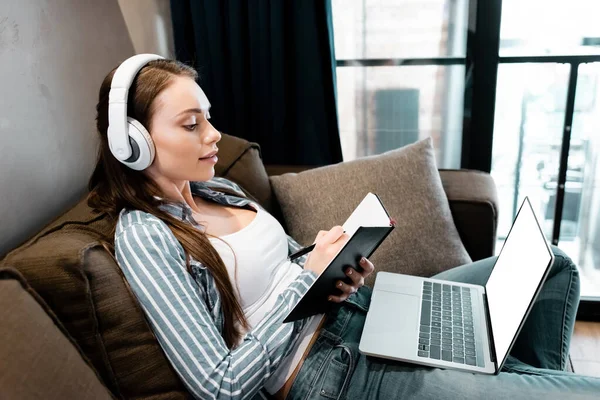 Mujer atractiva en auriculares inalámbricos escritura en portátil cerca de la computadora portátil con pantalla blanca, concepto de estudio en línea - foto de stock