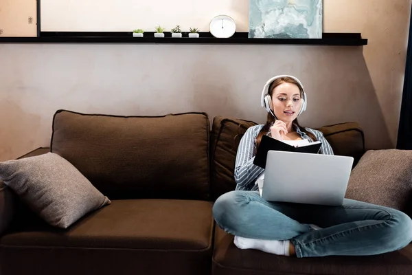 Mujer joven en auriculares inalámbricos mirando a la computadora portátil y la celebración de portátil con pluma en la sala de estar, concepto de estudio en línea - foto de stock