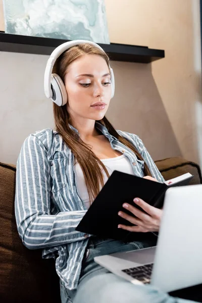 Selective focus of attractive woman in wireless headphones writing in notebook near laptop in living room, online study concept — Stock Photo