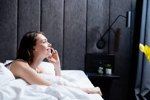 Seitenansicht einer glücklichen jungen Frau, die im Bett auf dem Smartphone spricht — Stockfoto