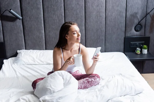 Atractiva mujer joven con teléfono inteligente y la celebración de la taza de café en la cama - foto de stock