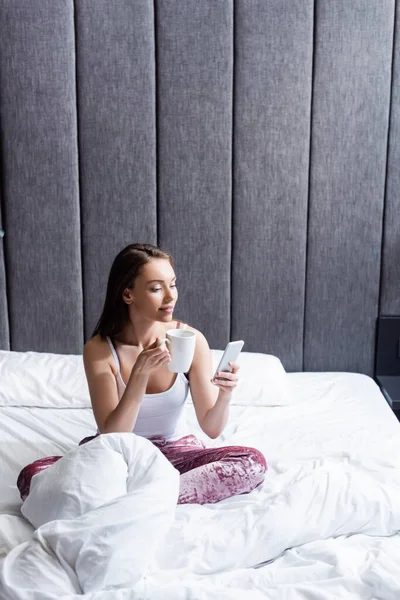 Beautiful young woman using smartphone and holding cup of coffee in bed — Stock Photo