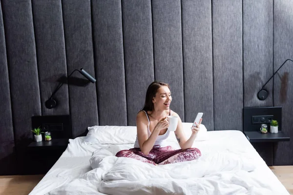 Happy young woman using smartphone and holding cup of coffee in bed — Stock Photo