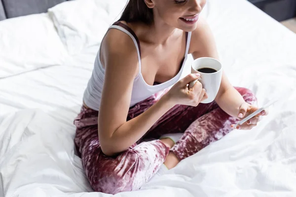 Vista recortada de la joven feliz usando el teléfono inteligente y la celebración de la taza de café en la cama - foto de stock