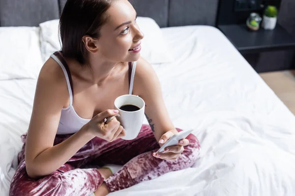 Happy young woman holding smartphone and cup of coffee in bed — Stock Photo