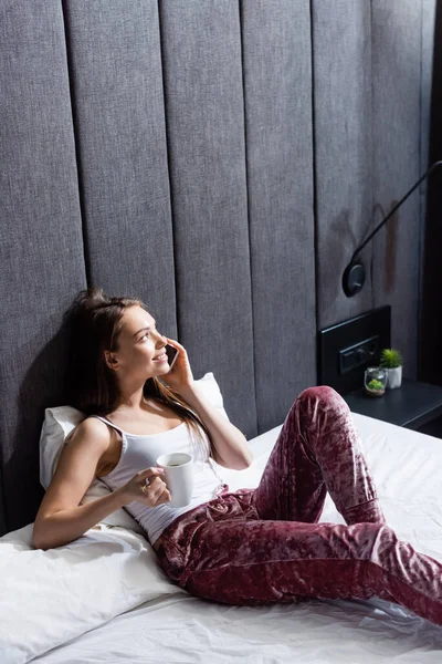 Mulher feliz falando no smartphone e segurando xícara de café na cama — Fotografia de Stock