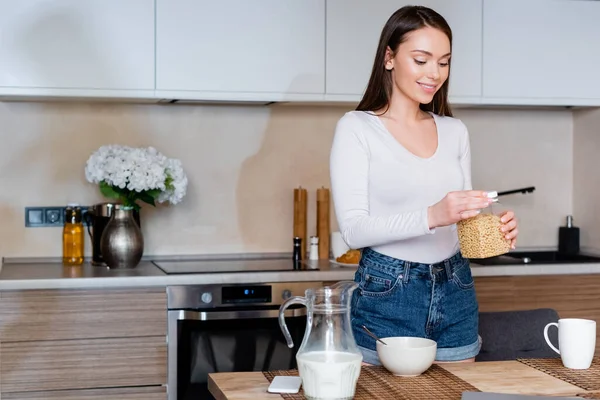 Fröhliches Mädchen hält Behälter mit Cornflakes in der Nähe von Schüssel und Krug mit Milch — Stockfoto