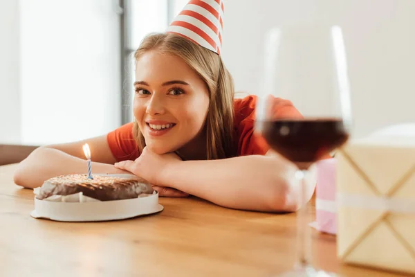 Foco seletivo da menina feliz na tampa do partido olhando para a câmera perto de bolo de aniversário, presentes e copo de vinho na mesa — Fotografia de Stock