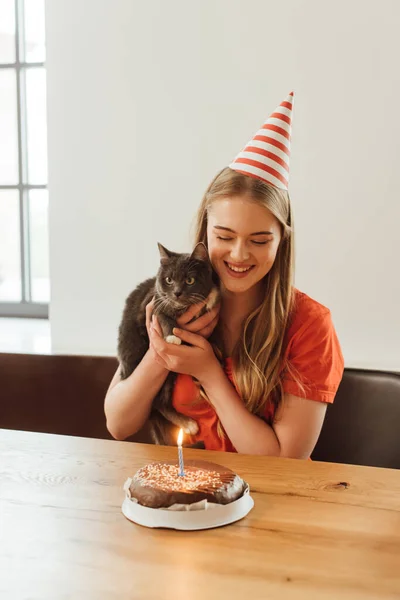 Happy girl in party cap looking at birthday cake and holding in arms cat — Stock Photo