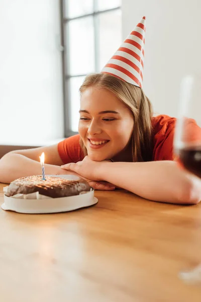 Selective focus of happy girl in party cap looking at burning candle on birthday cake — Stock Photo