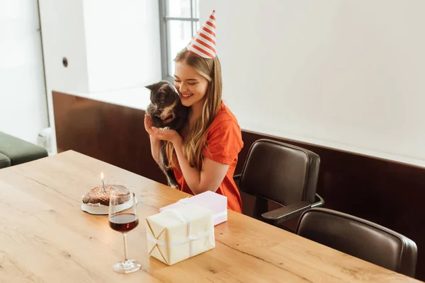 Щаслива дівчина у вечірній кепці тримає в руках кішку і дивиться на торт на день народження біля подарунків і червоне вино в склі — стокове фото