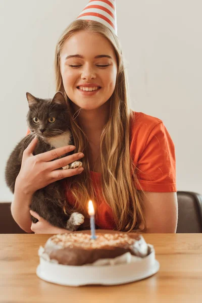 Enfoque selectivo de la chica feliz mirando la vela ardiente en pastel de cumpleaños y sosteniendo en brazos lindo gato - foto de stock