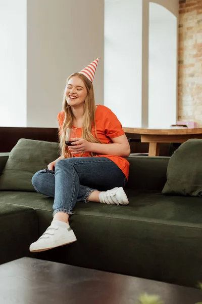 Foyer sélectif de la femme heureuse dans le chapeau de fête assis sur le canapé et tenant un verre de vin rouge — Photo de stock
