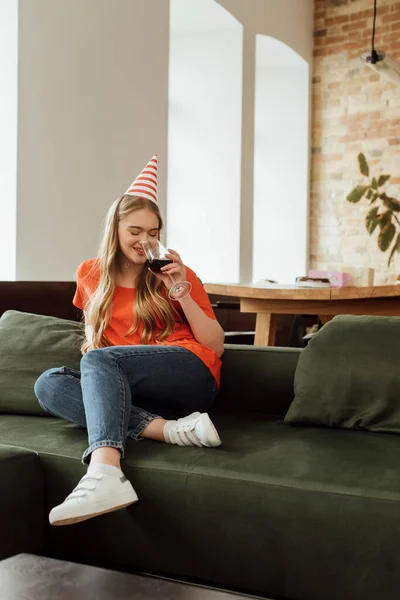 Счастливая молодая женщина в шапочке для вечеринок сидит на диване и пьет красное вино — стоковое фото