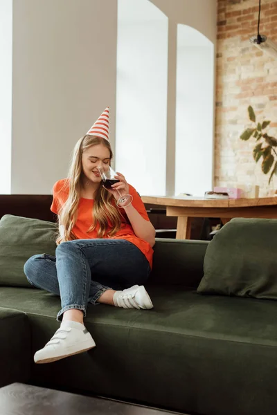 Happy young woman in party cap sitting on sofa and drinking red wine — Stock Photo
