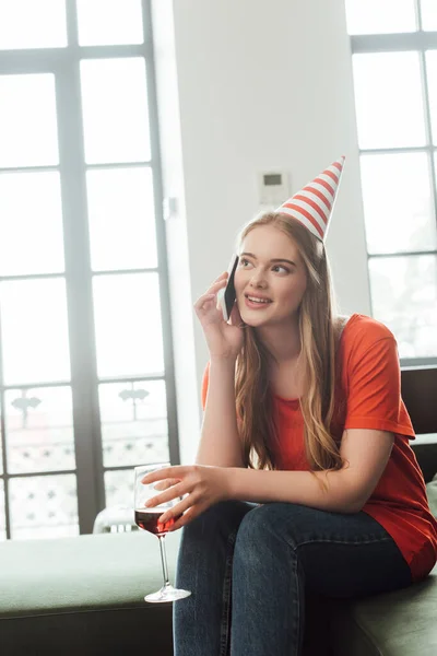 Fille heureuse dans le chapeau de fête tenant un verre de vin rouge et parlant sur smartphone à la maison — Photo de stock