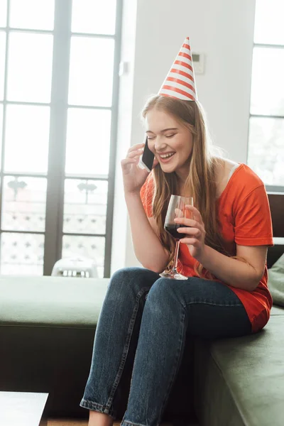 Chica sonriente en la tapa del partido sosteniendo una copa de vino tinto y hablando en el teléfono inteligente en casa - foto de stock