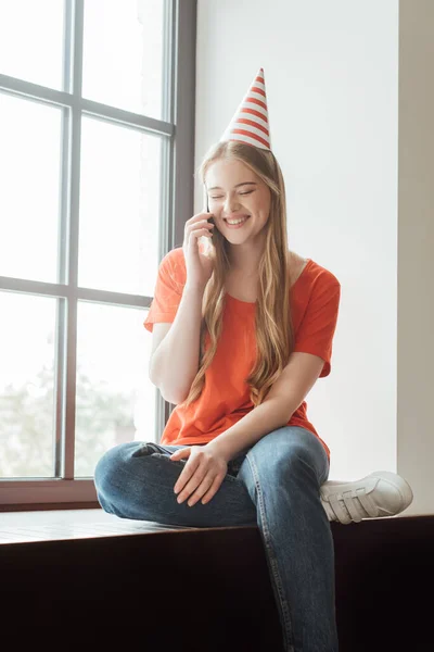 Positive girl in party cap sitting on window sill and talking on smartphone — Stock Photo