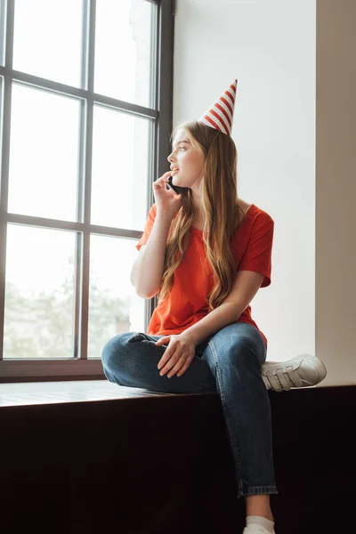 Attractive girl in party cap sitting on window sill and talking on smartphone while looking at window — Stock Photo
