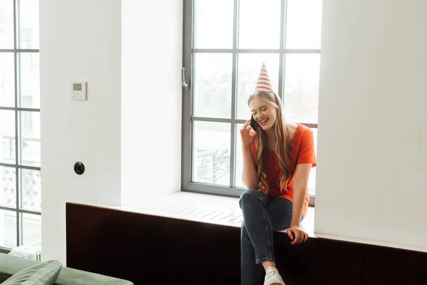 Chica sonriente en la gorra del partido sentado en el alféizar de la ventana y hablando en smartphone - foto de stock