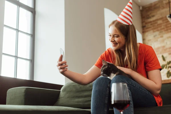 Enfoque selectivo de mujer joven atractiva en la tapa del partido tomando selfie con el gato cerca de la copa de vino tinto - foto de stock