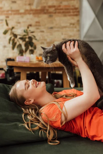 Cheerful woman lying on sofa and holding in arms cute cat — Stock Photo