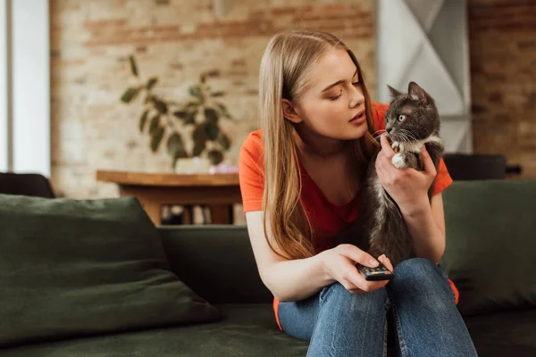 Junge Frau mit Fernbedienung und Blick auf süße Katze — Stockfoto