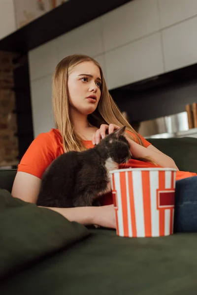 Messa a fuoco selettiva di giovane donna toccando gatto mentre guarda film vicino secchio popcorn — Foto stock