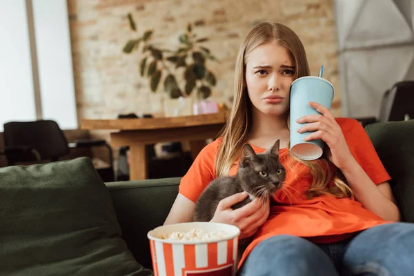 Selective focus of sad woman holding disposable cup with soda near popcorn bucket and cat while watching movie in living room — Stock Photo