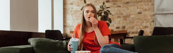 Panoramic shot of attractive girl eating popcorn, holding disposable cup and watching movie in living room — Stock Photo